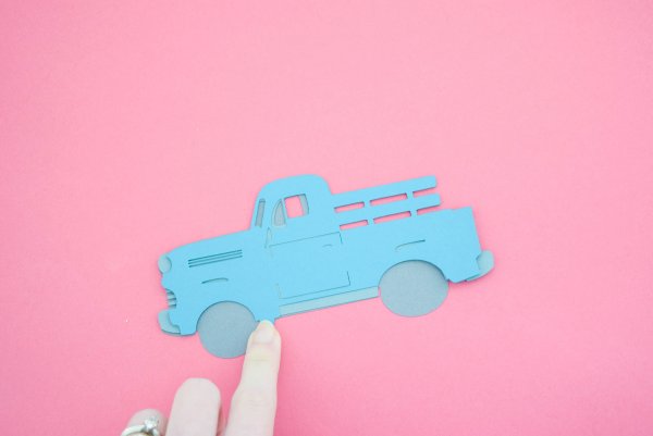 Abbi Kirsten's finger rests on the vintage Christmas truck ornament in it's early stages. The three truck pieces have been layered together and lay on a pink paper background. These parts can be cut out using a Cricut cutting machine and a free SVG file.