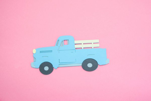 The 3D layered paper vintage truck ornament, complete with tires and a rack, rests on pink paper. This truck will look great on any Christmas tree and is a fun project for kids, too. 