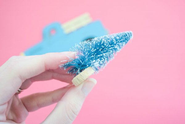 In a close-up, Abbi demonstrates how to take a miniature bottle brush Christmas tree and bend the base at a 90 degree angle. In the background is the near-completed vintage truck ornament and the pink backdrop for this craft. 