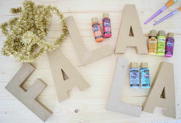 A wooden table full of supplies needed to make DIY decorative tinsel Christmas letters. Supplies include gold tinsel in the corner, above the carboard letters spelling out "Fa La La." Craft paint bottles and brushes round out the supply list. 