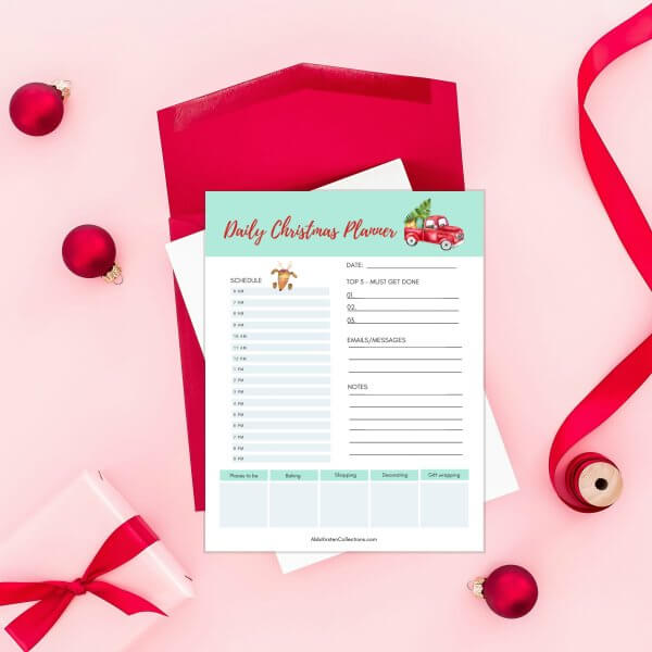 A holiday Christmas planner printable form lays on a red envelope on a pink table with red ribbon amd tiny red ornaments scattered around for decoration. 