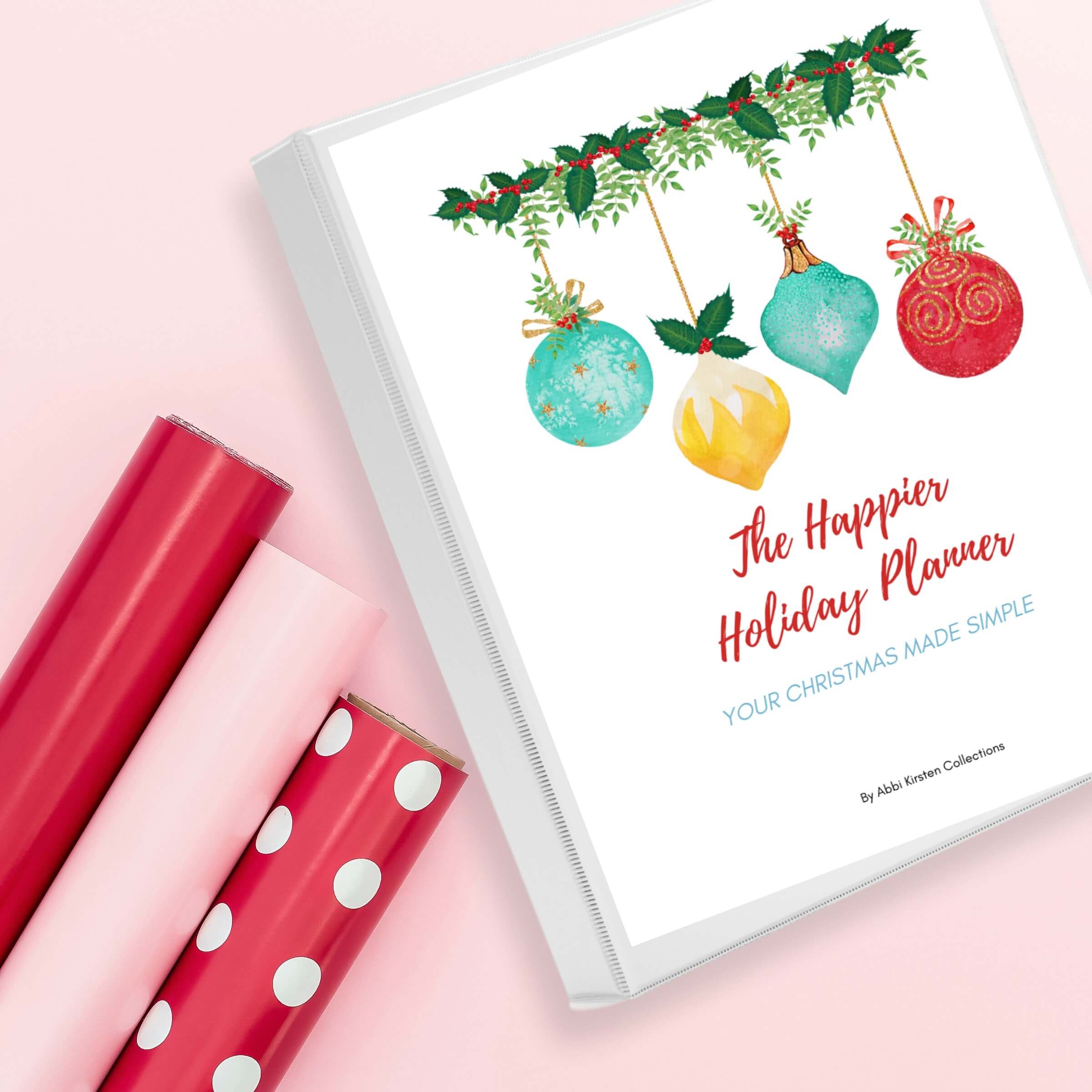 Flat lay photo of The Happier Holiday Planner on a pink surface beside three gift wrapping paper