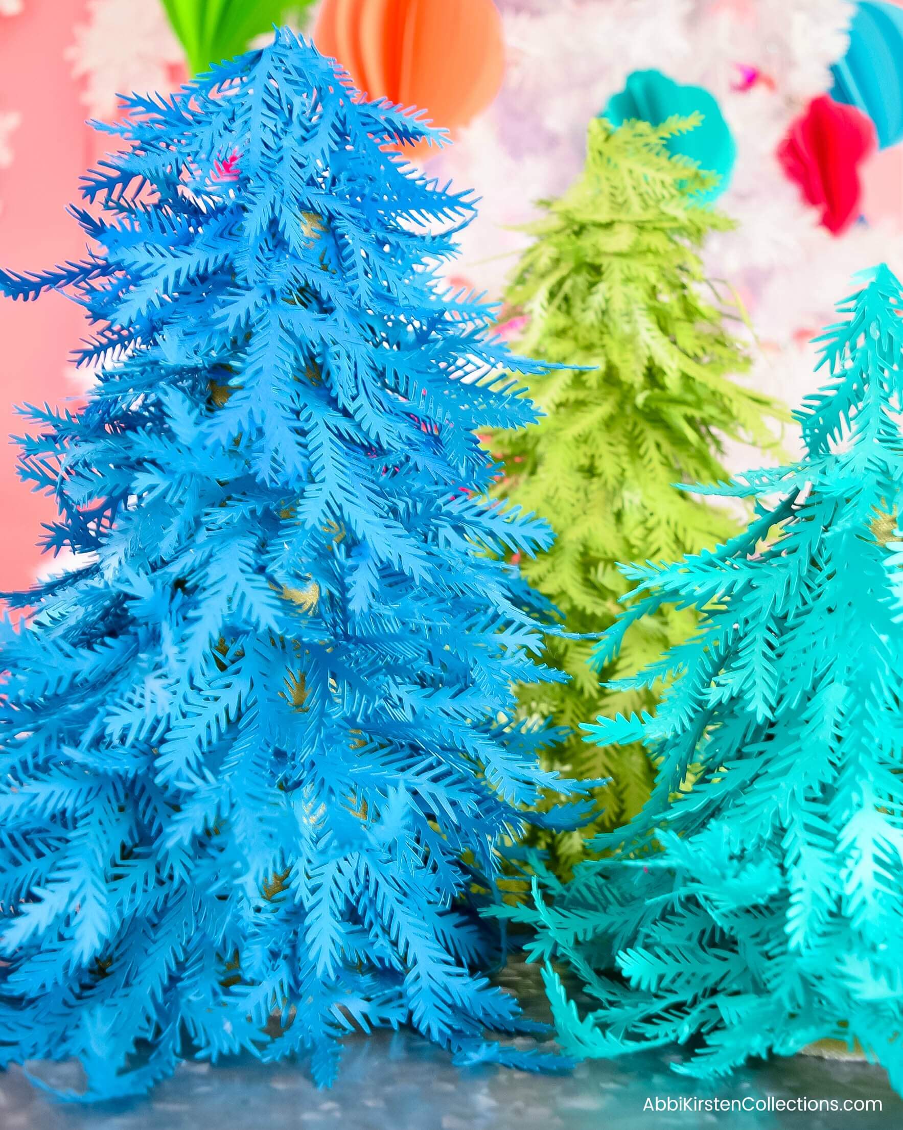 Three colorful paper Christmas trees made with blue, teal, and green paper stand in front of a festive background.