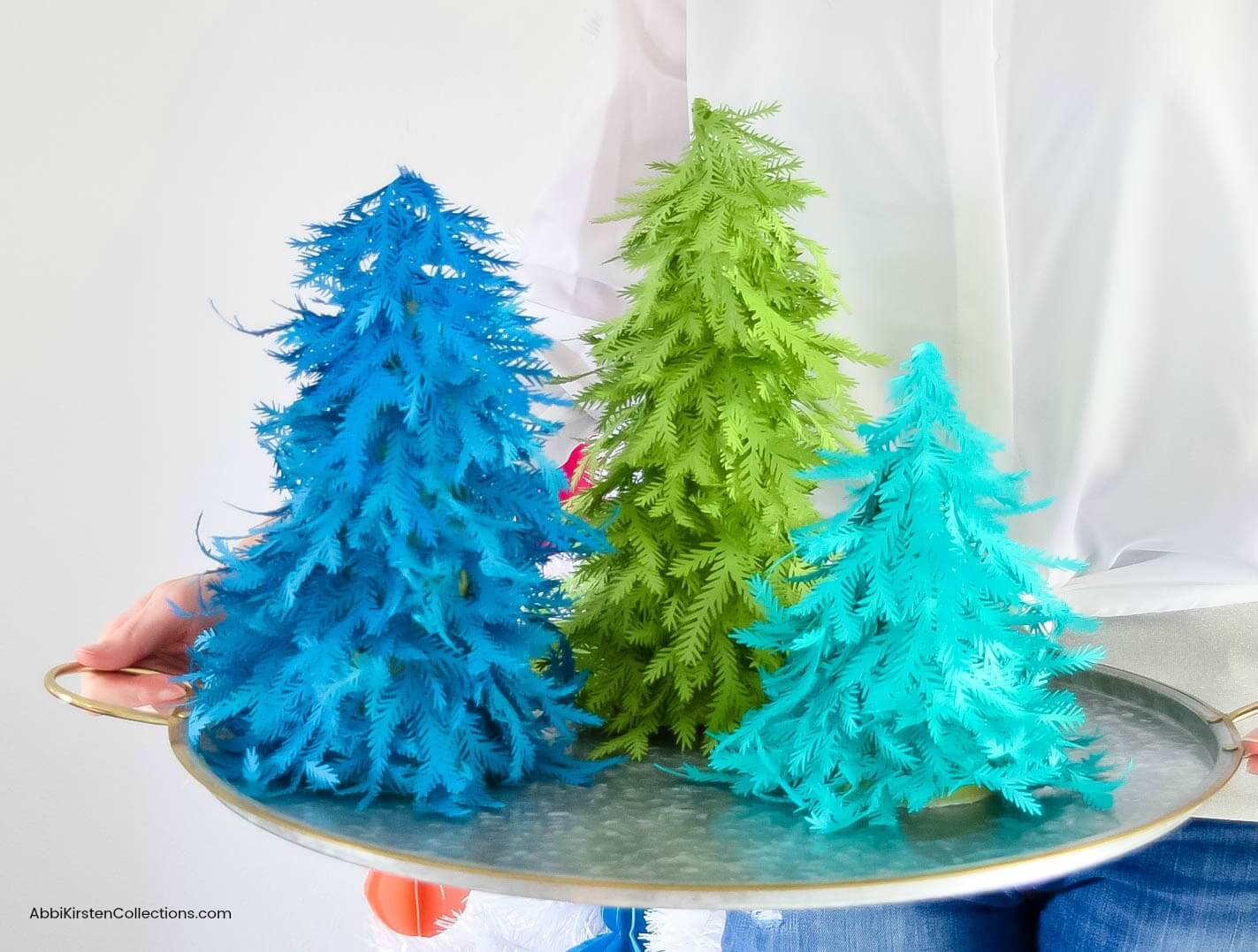 Three small paper Christmas trees of different sizes, sitting on a metal tray. The tallest paper tree is made with green paper, the middle tree is blue, and the smallest tree is a light teal color.