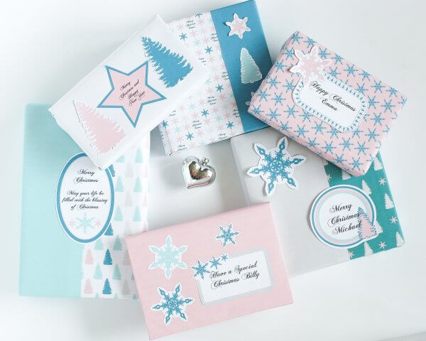 An overhead view of six presents stacked in a circle with a silver heart bobble in the center. The presents are on an off-white backdrop and are wrapped in custom pink, white, blue and teal-colored wrapping paper. 