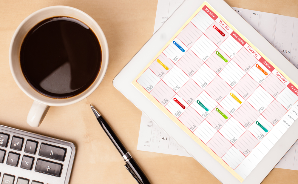 An overhead image of a workspace with a color-coded paper calendar, computer keyboard, and a mug of black coffee. Using an organized calendar is a simple and effective time management tip for craft business owners.