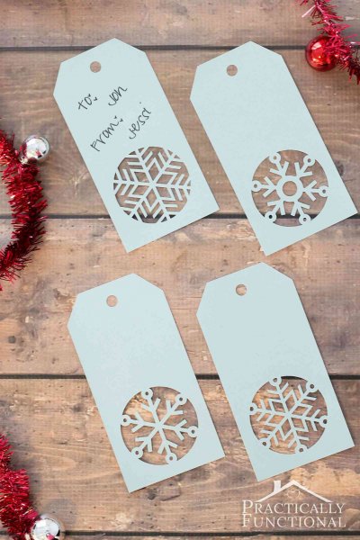 Four printable gift tags each have an intricate paper snowflake cut into the bottom of the tag. The tags lay flat on a wooden table, surrounded by red garland. 
