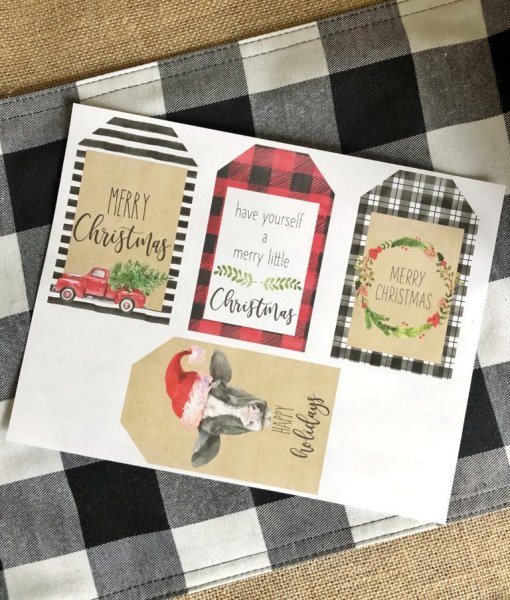 Rustic paper gift tags for Christmas or holiday presents have been printed, four to a page, and laid on a black and white checked linen. Each gift tag has a unique pattern – a buffalo check pattern in red, black, and white, black and white stripes, and a cartoon Christmas cow.