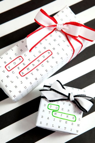 Two gifts wrapped in a customizable word-search game gift wrap. The top present has the words "For" and "Amanda" circled in red, and the bottom present has "To" and "Mike" circled in green. The presents sit on a black and white-striped surface. Make gift-giving a fun game!