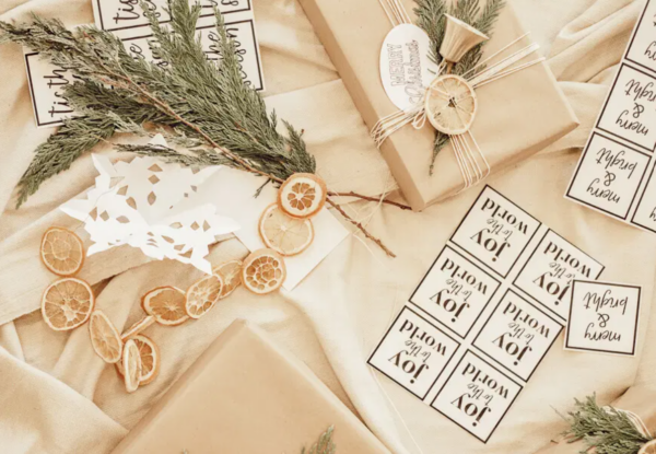Sheets of free printable Christmas gift tags sit next to Scandinavian-wrapped holiday gifts in tan paper and light tan twine with dried orange slices as decorations. The gift tags say “joy to the world” and “‘tis the season.”