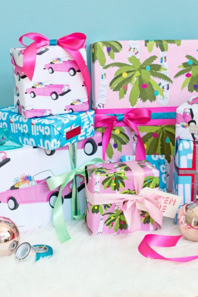 Multiple Christmas presents wrapped in bright pastel and beach-themed wrapping paper. The illustrations on the paper include a pink Cadillac and tropical trees with Christmas lights. 