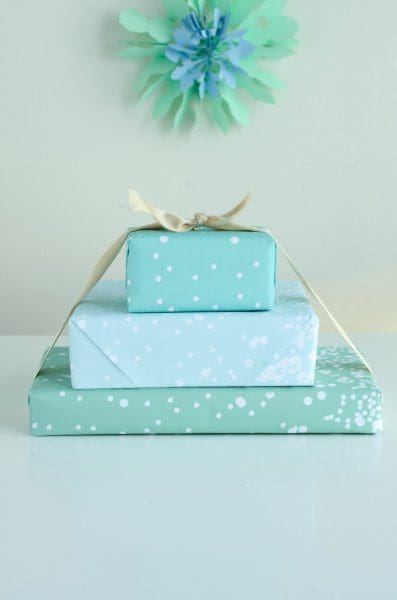 Three blue presents, tied with a yellow ribbon, are sitting on a blue tabletop in front of a blue-green wall. The printable paper is Tiffany-blue with dots of white snow.