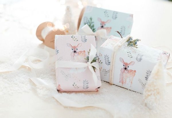 A pile of presents rests on white blankets. These presents are wrapped in pale blue and white wrapping paper with illustrations of a delicate pastel deer, and wrapped with white ribbon. 