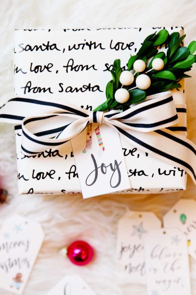 A close-up image of a square present wrapped in white paper with black handwriting that says "With love, from Santa" repeatedly. The present is finished with a black and white ribbon and a sprig of holly. The printed gift tag has black script that reads "Joy."