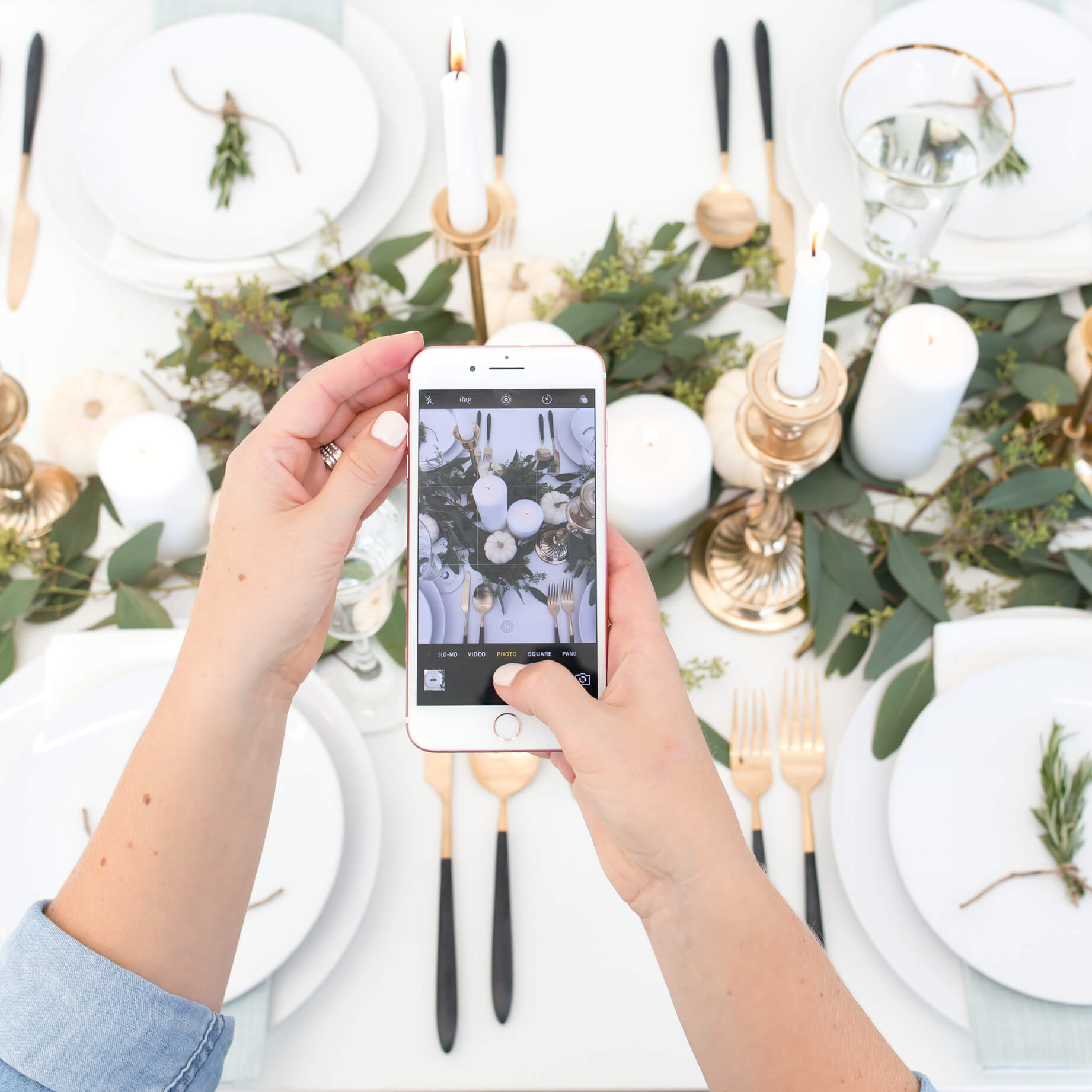A woman uses her smartphone to take a picture of a beautifully curated holiday tablescape. White dinner plates, gold and black silverware, and gold candlesticks are set up around a greenery centerpiece. 