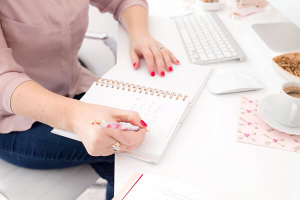 A woman sits at a desk in front of a computer keyboard writing in her planner. A cup of coffee is nearby. Writing goals out is a great time management tip. 