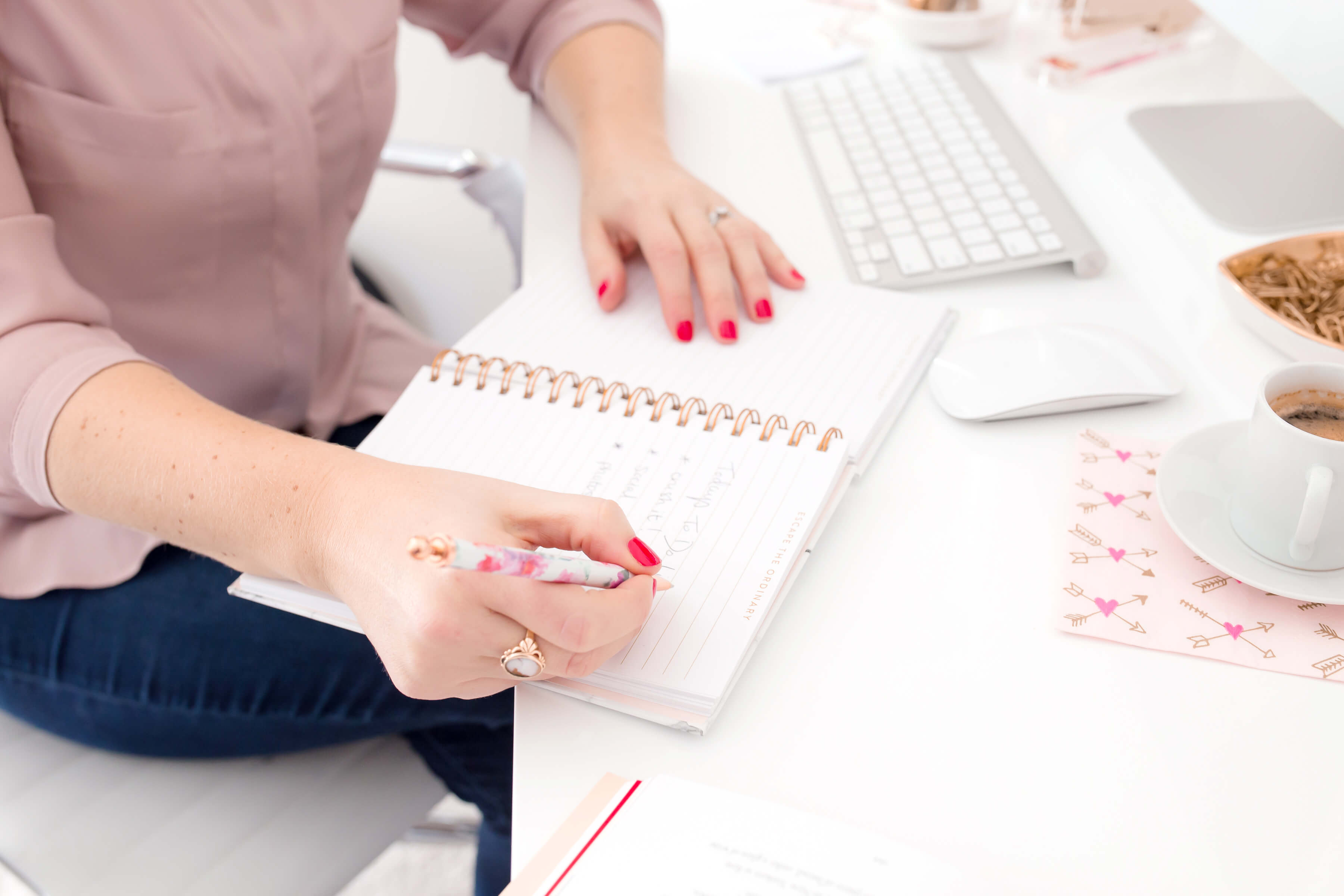 A woman sits at a white desk, working on writing down her business goals in a planner. There’s a computer and mug of coffee next to her on the desk.