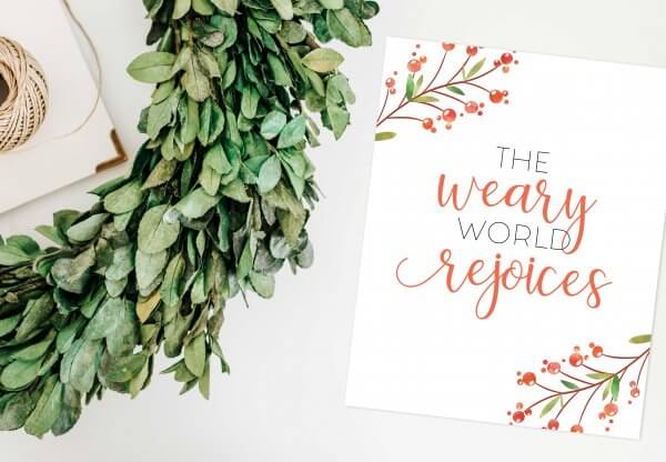 A half-cirlce of garland lays next to a handmade Christmas card that reads "The weary world rejoices."