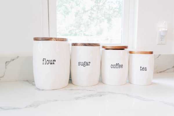 Four white canisters with woodgrain tops set on a marble countertop below a window. Each canister is labeled "flour, sugar, coffee, tea, respectively.