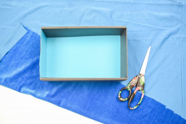 An empty shoe box on top of light blue velvet fabric and a pair of scissors. This easy step-by-step fabric storage box is an easy craft for kids and adults. 