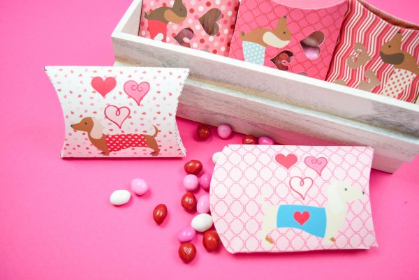 A pink tabletop holds adorable dog-themed Valentine's Day candy boxes in holiday colors. A wooden box contains even more candy boxes. Candy is spilling out onto the table. 