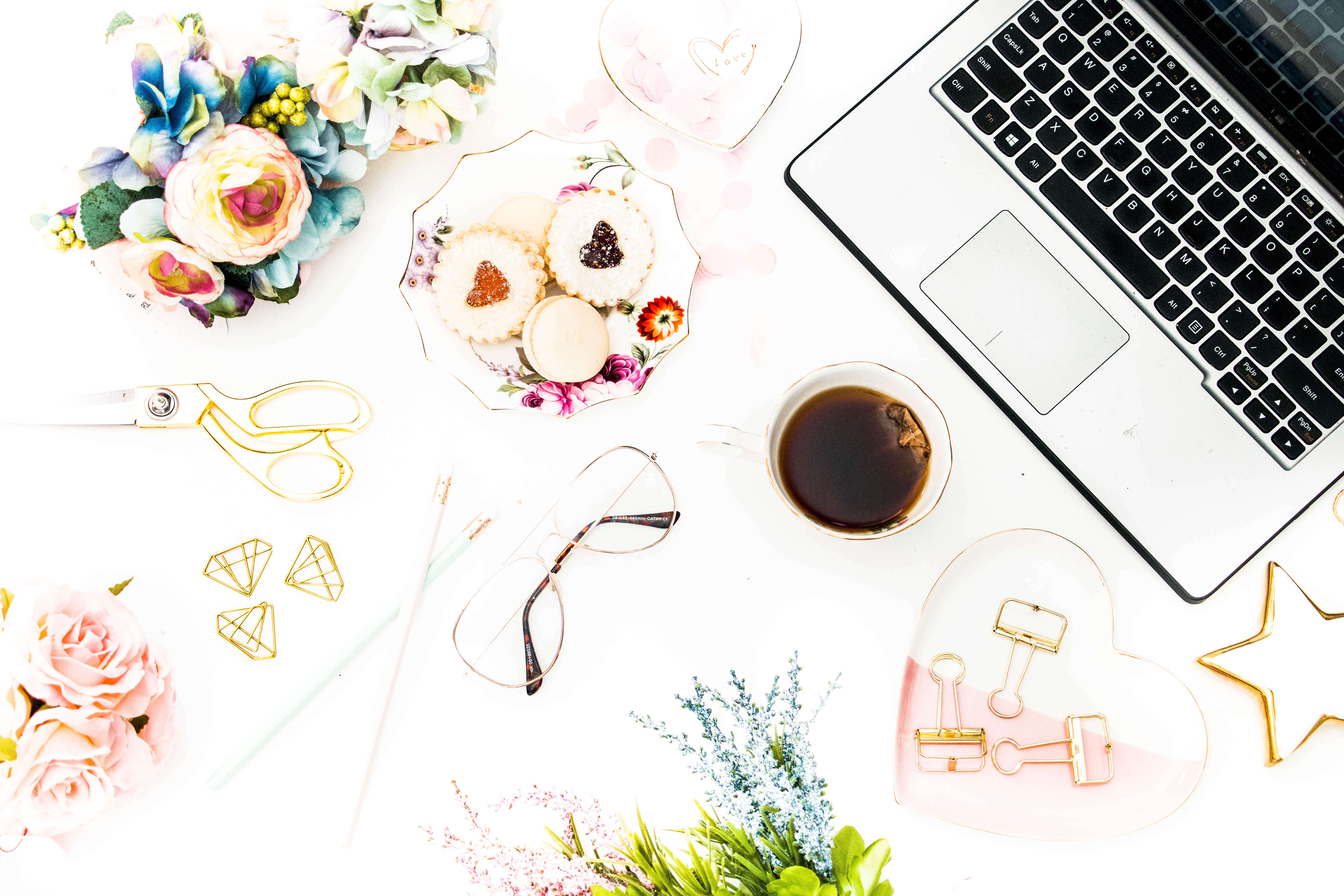 A desk with multiple pastel colored and gold items, biscuits and tea, and a silver computer. 