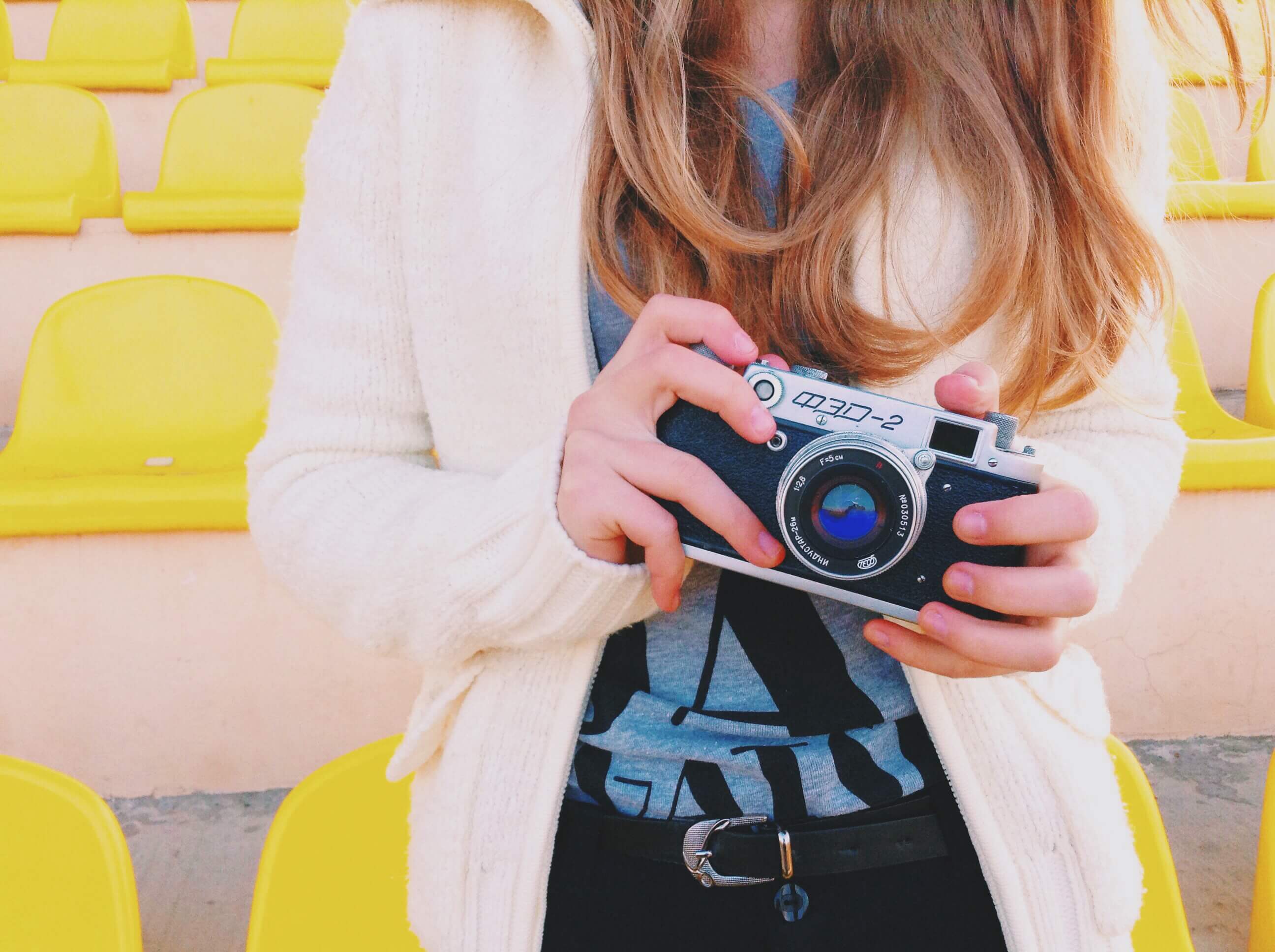 A woman with long blonde hair holds a vintage film camera in her hands. She's wearing a white sweater over a gray t-shirt with jeans.