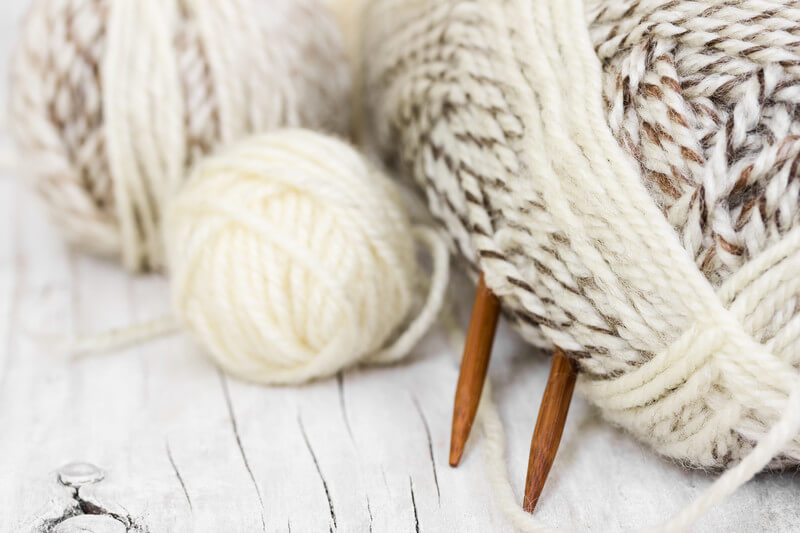A close-up image of cream and tan colored balls of yarn and wooden knitting needles on a white wooden table. 