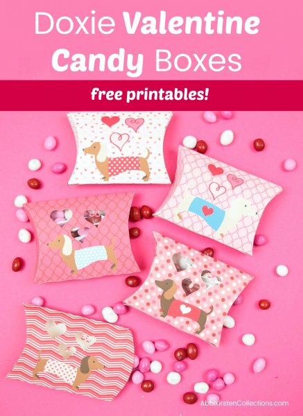 Daschund-themed Valentine's Day pillow boxes lay on a pink background. Holiday-colored candy spills out. The text says "Doxie valentine candy boxes free printables!"