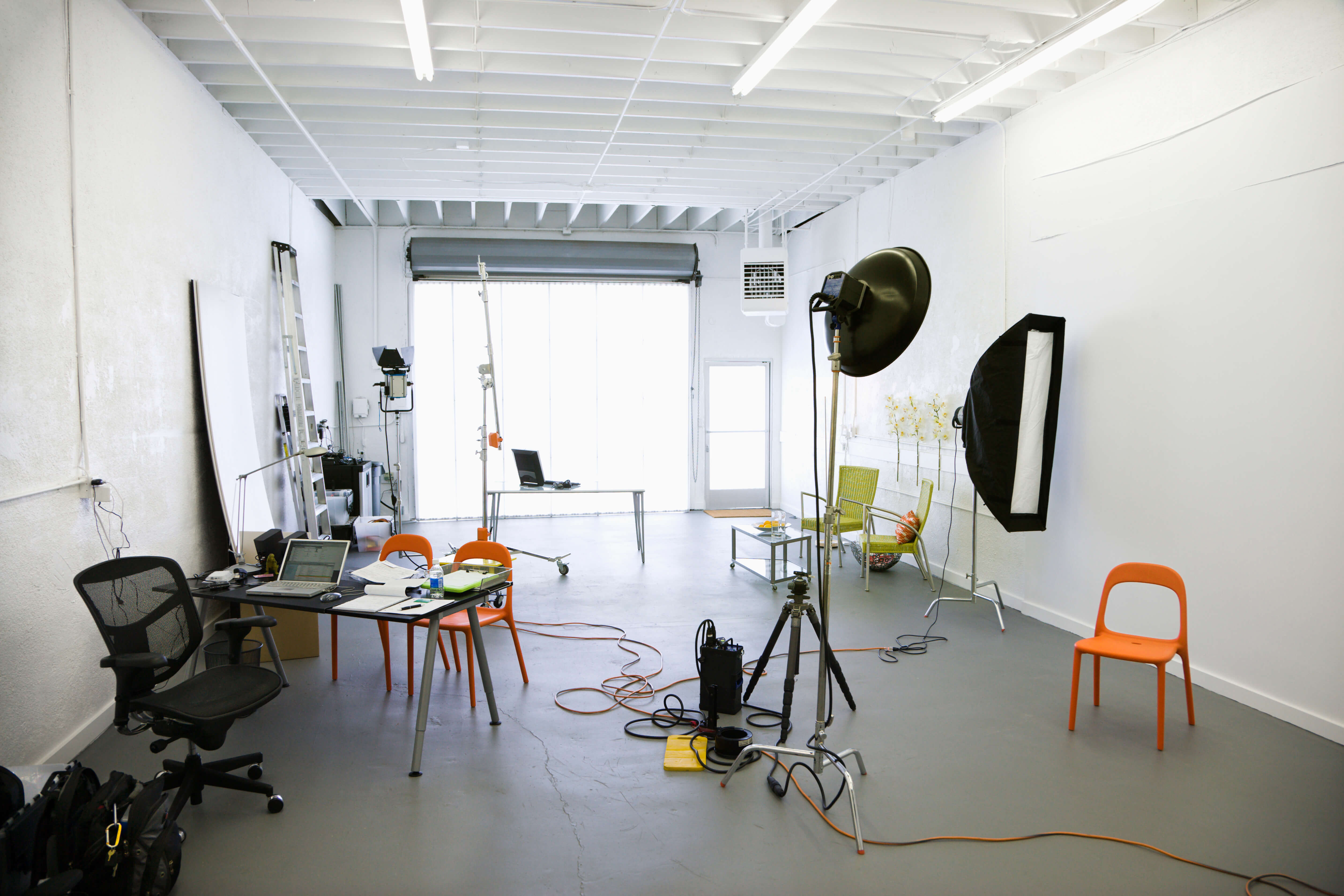 An all-white photography studio is set up with photography equipment, a small work area with a desk, chair, and laptop, photo space, and a large floor-to-ceiling opening letting in natural light.