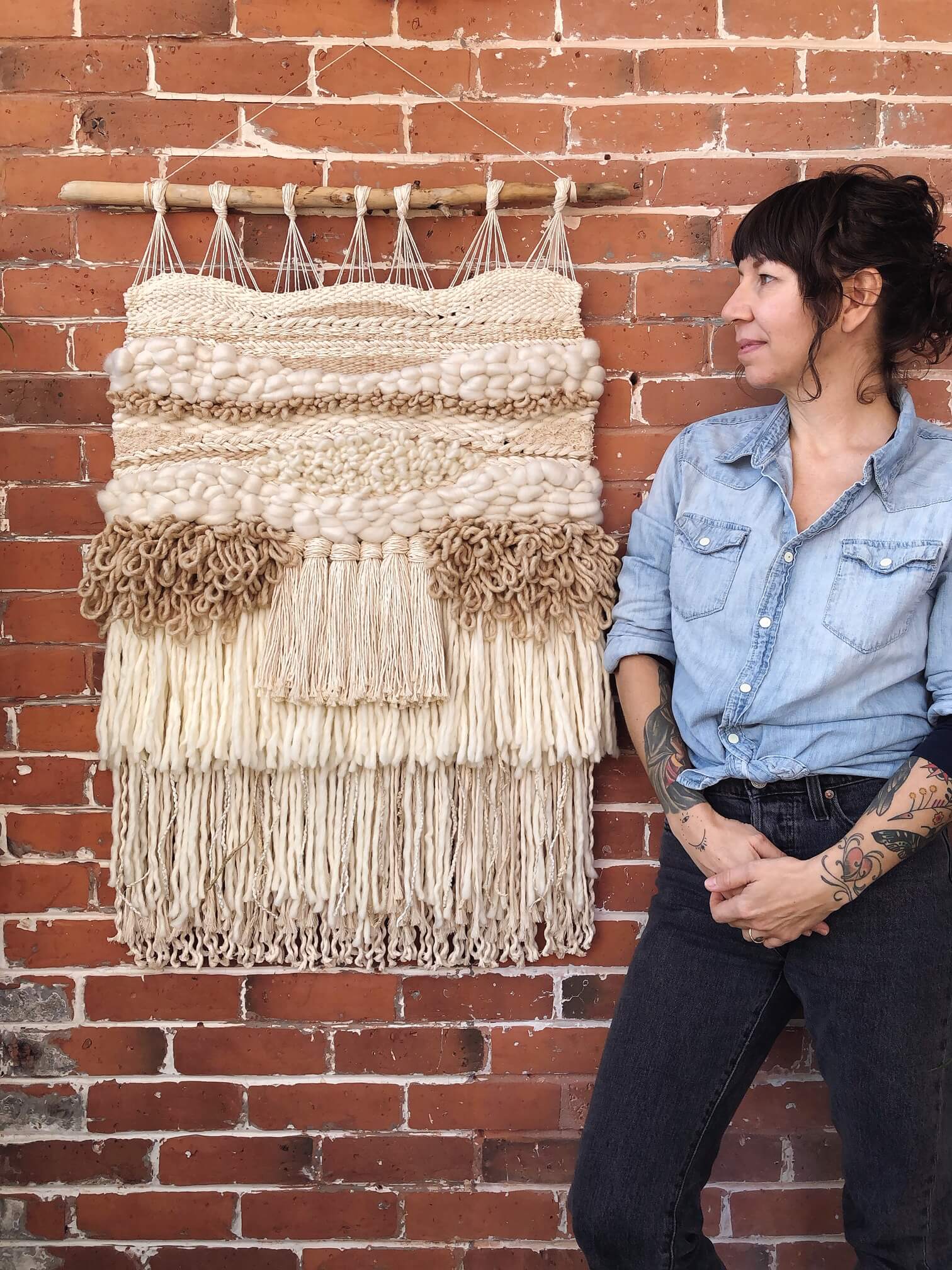 A young woman with dark hair poses next to a large macrame wall hanging mounted on a brick wall. This macrame wall art has a neutral color palette with tan, cream, and light brown threads intricately woven together.