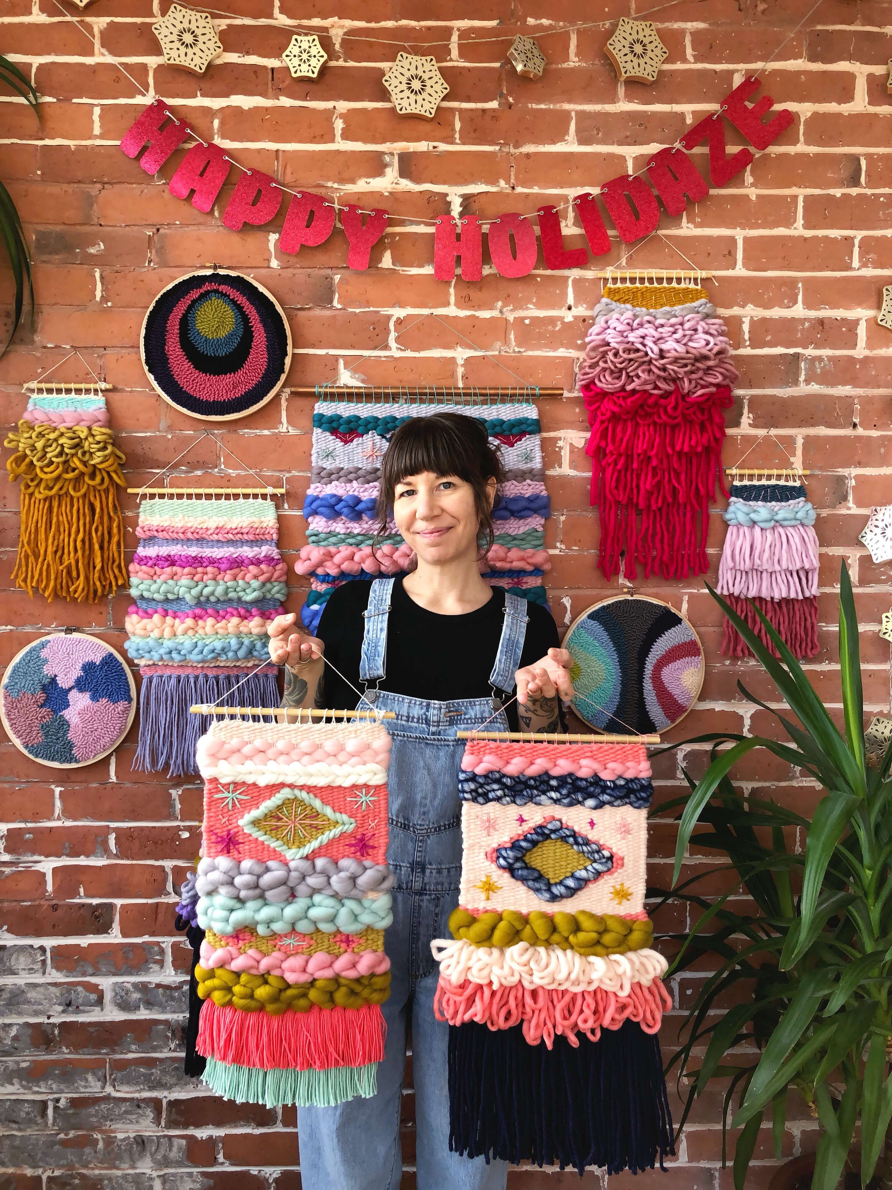 A woman poses with her macrame wall hangings of different sizes and color schemes. She's holding one wall hanging in each hand, and stands in front of a brick wall where several other macrame wall hangings of various shapes and sizes hang.