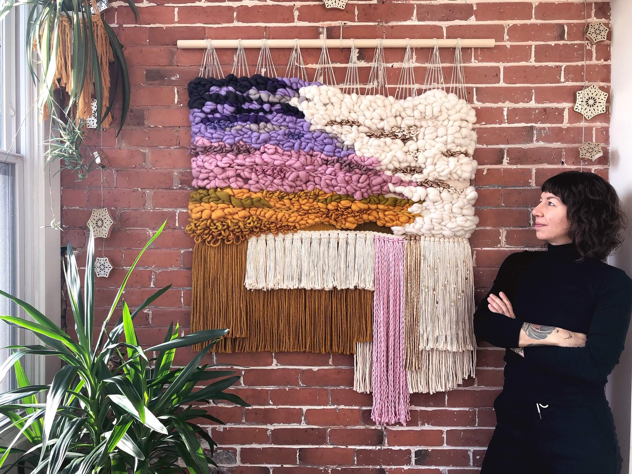 A woman with short black hair, dressed in all black, poses next to a large macrame wall hanging that's hung on a brick wall. The wall hanging features intricately woven yarn in many colors like brown, yellow, shades of pink, purple, and cream.