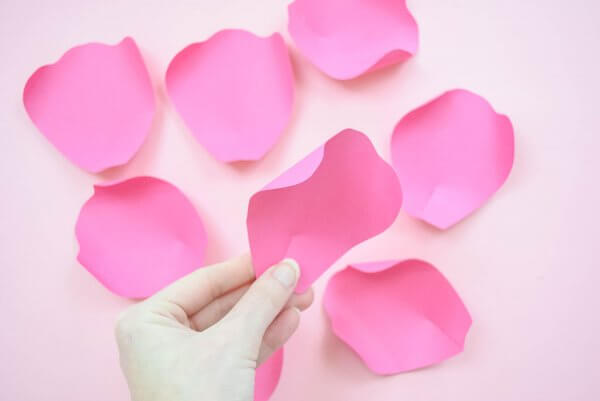 Small, curled pink paper petals lay on a tabletop ready for assembly. Abbi Kirsten's hands hold a finished small petal above the others. 