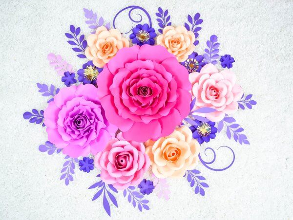 This paper rose tutorial by Abbi Kirsten makes this Penelope rose paper flower. A bunch of pink, purple, and orange paper flowers of varying sizes are bundled and sit on a bed of blue foliage. The overall look is stunning and breathtakingly beautiful. 