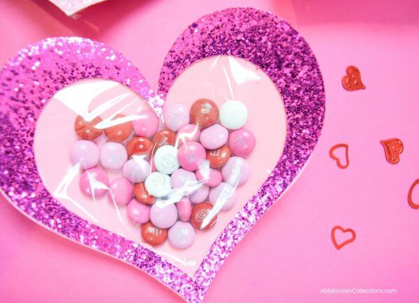 A single candy-filled Valentine's Day heart made from purple glitter cardstock lays on a pink backdrop. These easy Valentine's Day crafts include templates and a tutorial.  