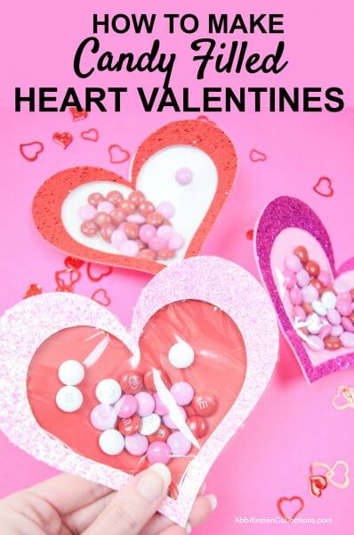 Paper heart envelopes made with glitter paper and a cellophane window showcases Valentine's Day candy. These hearts lay on a pink background with text that reads "How to make candy filled heart valentines."