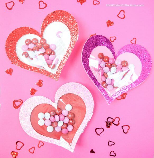 Three glitter paper hearts with cellophane windows lay on a pink backdrop with red heart confetti. These hearts are filled with candy for your Galentine's Day! 