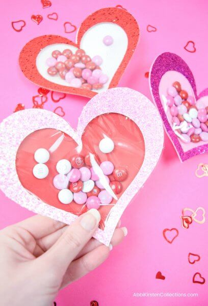 Abbi holds a heart-shaped candy-filled paper valentine. Other colors of hearts are in the background on a pink tabletop. Easy Valentine's Craft with Free SVG and Tutorial.