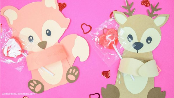 An paper orange fox and tan squirrel are hugging their candy heart suckers while laying on a pink backdrop surrounded by a sprinkle of red heart confetti. The fox woodland Valentine candy hugger template is free from Abbi Kirsten's Freebie vault. 