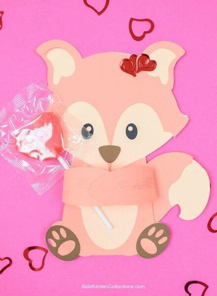 Light-brown fox candy hugger holding a red heart-shaped candy lollipop. Red cutout hearts surround the papercraft which rests on a dark pink paper backdrop. You can make this Valentine animal with Abbi Kirsten's instructions and free SVG cut file. 