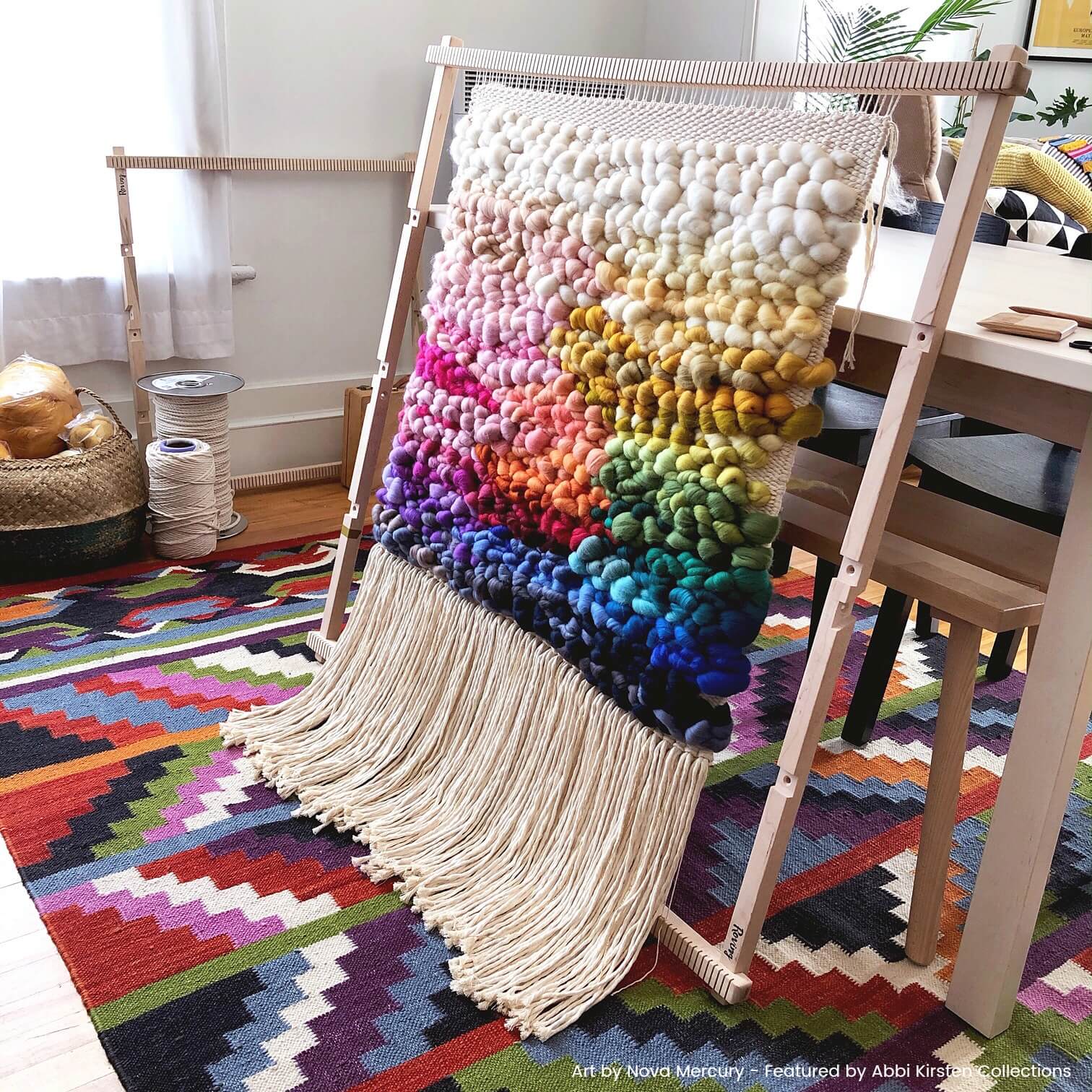 An in-progress macrame wall hanging hangs from a weaving loom while being made in a home craft room. This wall hanging features a rainbow patters, with colorful threads ranging from whites and creams to rich shades of blue and gray.