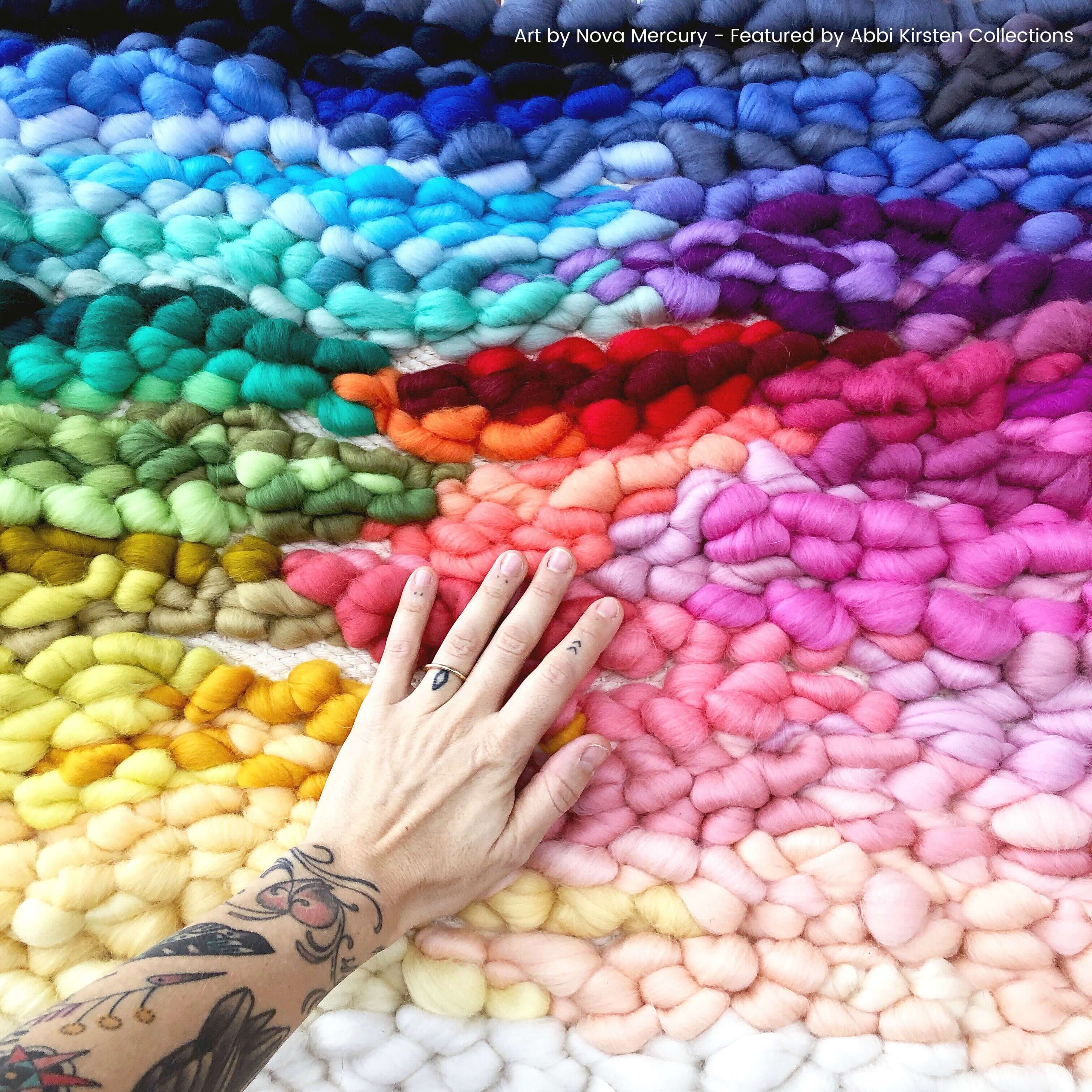 A woman's hand and tattooed arm rest on a rainbow macrame wall hanging made with colorful threads in shades of blue, green, red, orange, yellow, pinks, and purples.