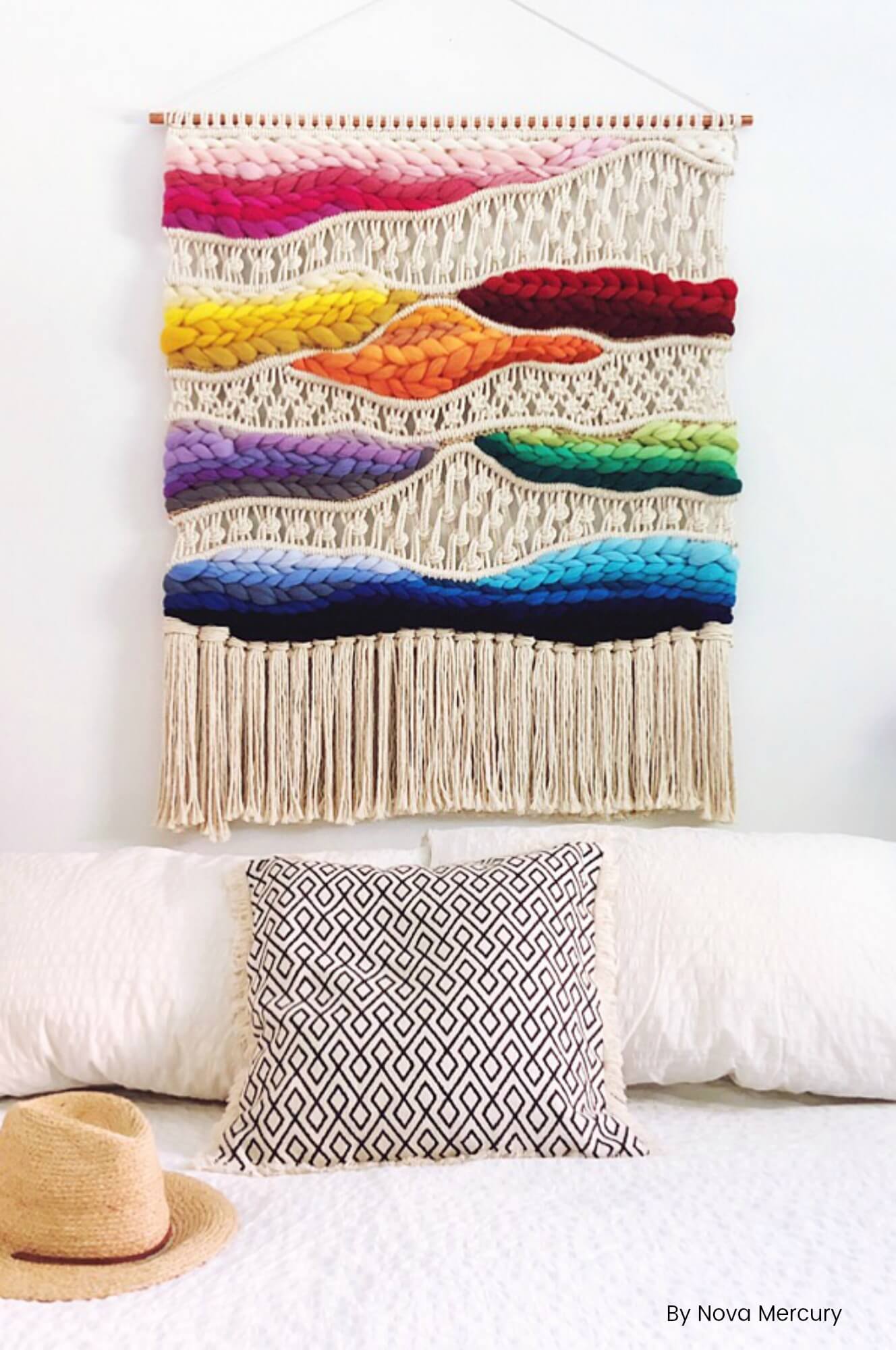 A large macrame wall hanging hangs over a white bed. The wall hanging is made with tan yarn with blocks of bright primary rainbow colors.