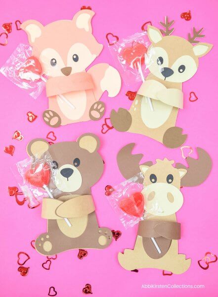 Four paper woodland animals are viewed from overhead on a dark pink background with red heart confetti strewn around them. In a variety of brown colors, each hugger hold a red heart candy lollipop. The cute creatures were cut out using a Cricut machine or scissors and include a fox, a squirrel, a bear and a moose
