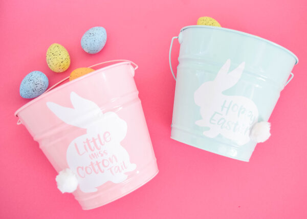 Two metal buckets - one light pink, one light blue - white white easter bunny vinyl cutouts attached, sit against a bright pink background. Yellow and blue easter eggs spill out from the tops of the buckets.