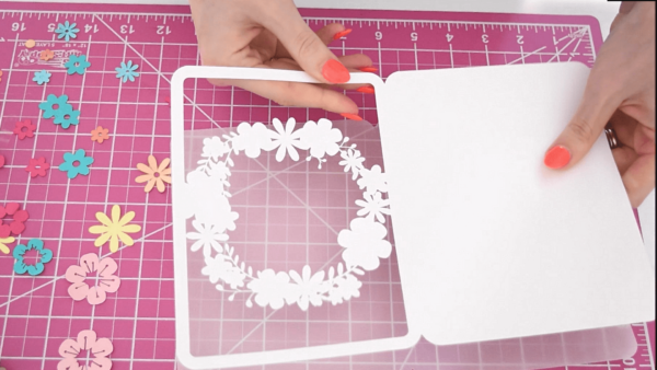 A white base for a DIY floral Mother's Day card alongside pastel flower cutouts on a pink workspace. 