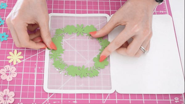 A green wreath being glued onto a white card atop a hot pink workspace. 
