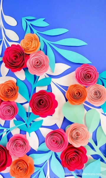 Groups of paper rosette flowers made with pink, red, and orange paper, and while and blue paper vine stems sit on a blue background.