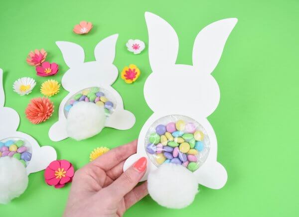 Abbi's hand holds one candy filled Easter Bunny craft. Two more, filled with pastel candy, lay on a light green backdrop surrounded by yellow, orange and white spring flowers. Free templates included!
